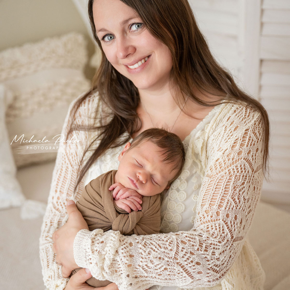 Mama hält ihr schlafendes Neugeborenes eng im Arm und lächelt warm in die Kamera.